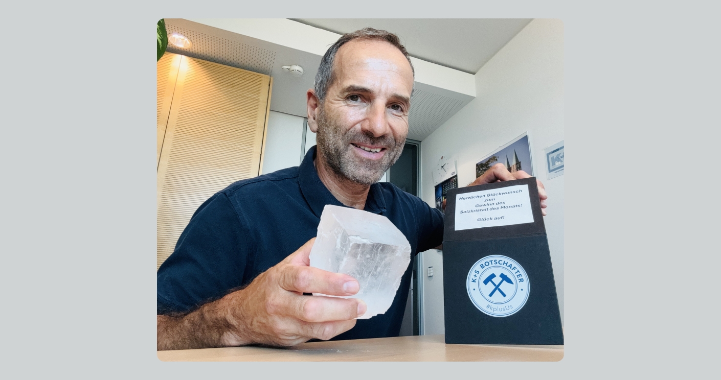 Aufnahme eines lächelnden Mannes am Schreibtisch eines modernen Büros. Er schaut in die Kamera, hält in der einen Hand einen Salzkristall, in der anderen eine Urkunde, auf der steht: Herzlichen Glückwunsch zum Gewinn des Salzkristall des Monats! Glück auf!
