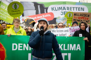 Luisa Neubauer in schwarzer Jacke, mit Käppi und Mikrofon bei einem Protest für Klimaschutzmaßnahmen vor dem Konrad-Adenauer-Haus in Berlin.. Dahinter sind Aktivist:innen mit einem großen Banner und Plakaten zu sehen.