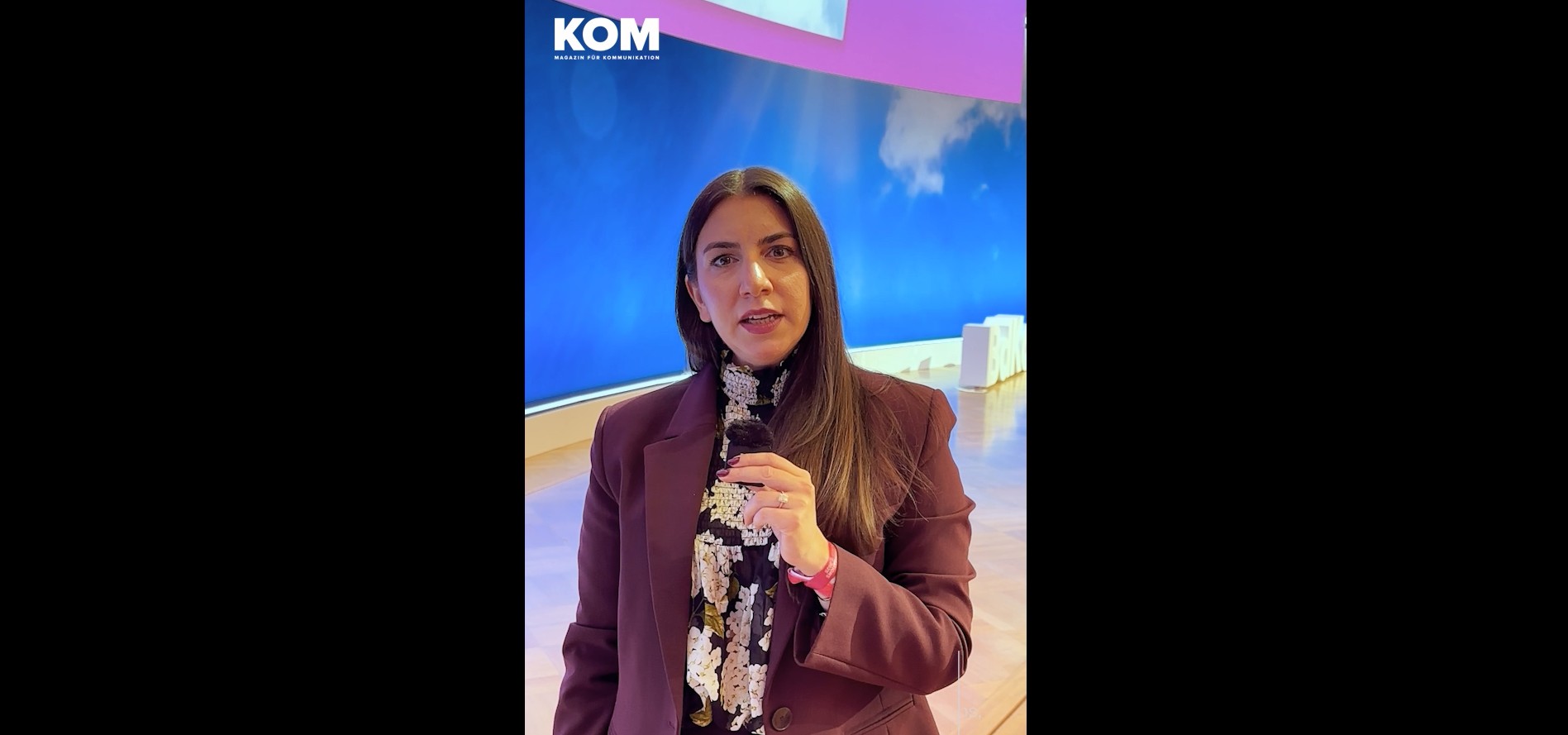 Frau mit langen, dunklen Haaren spricht in ein Mikrofon vor einem blauen Hintergrund. Sie trägt einen Bluse mit auffälligem Blumenmuster und einen Blazer in dunklem Altrosé.