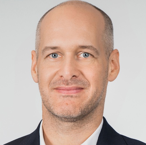 Porträt eines Mannes mit glattem Kopf, blauen Augen und leichtem Bart, lächelnd vor neutralem Hintergrund.