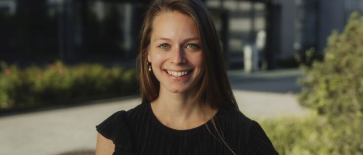 Porträt einer lächelnden Frau mit langen Haaren, die ein schwarzes Oberteil trägt, vor einer modernen Gebäudeumgebung.