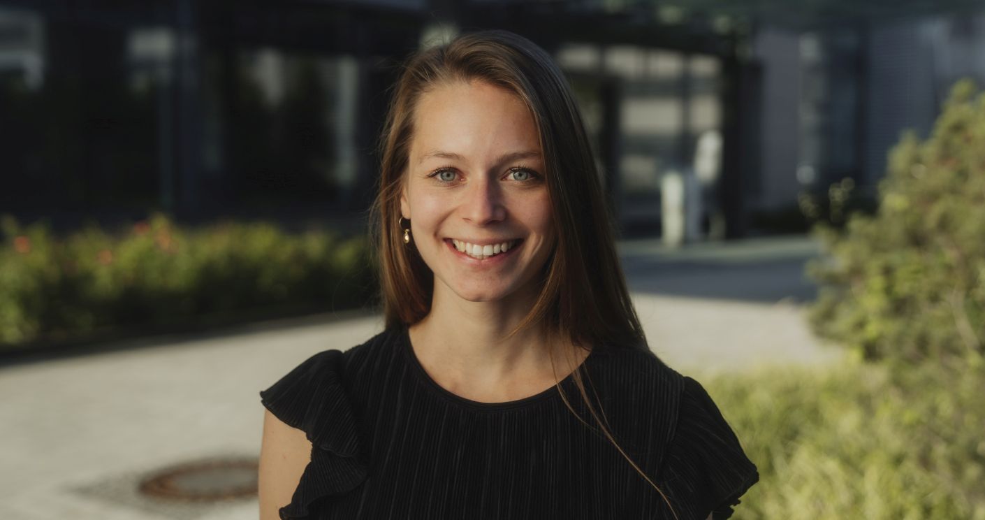 Porträt einer lächelnden Frau mit langen Haaren, die ein schwarzes Oberteil trägt, vor einer modernen Gebäudeumgebung.