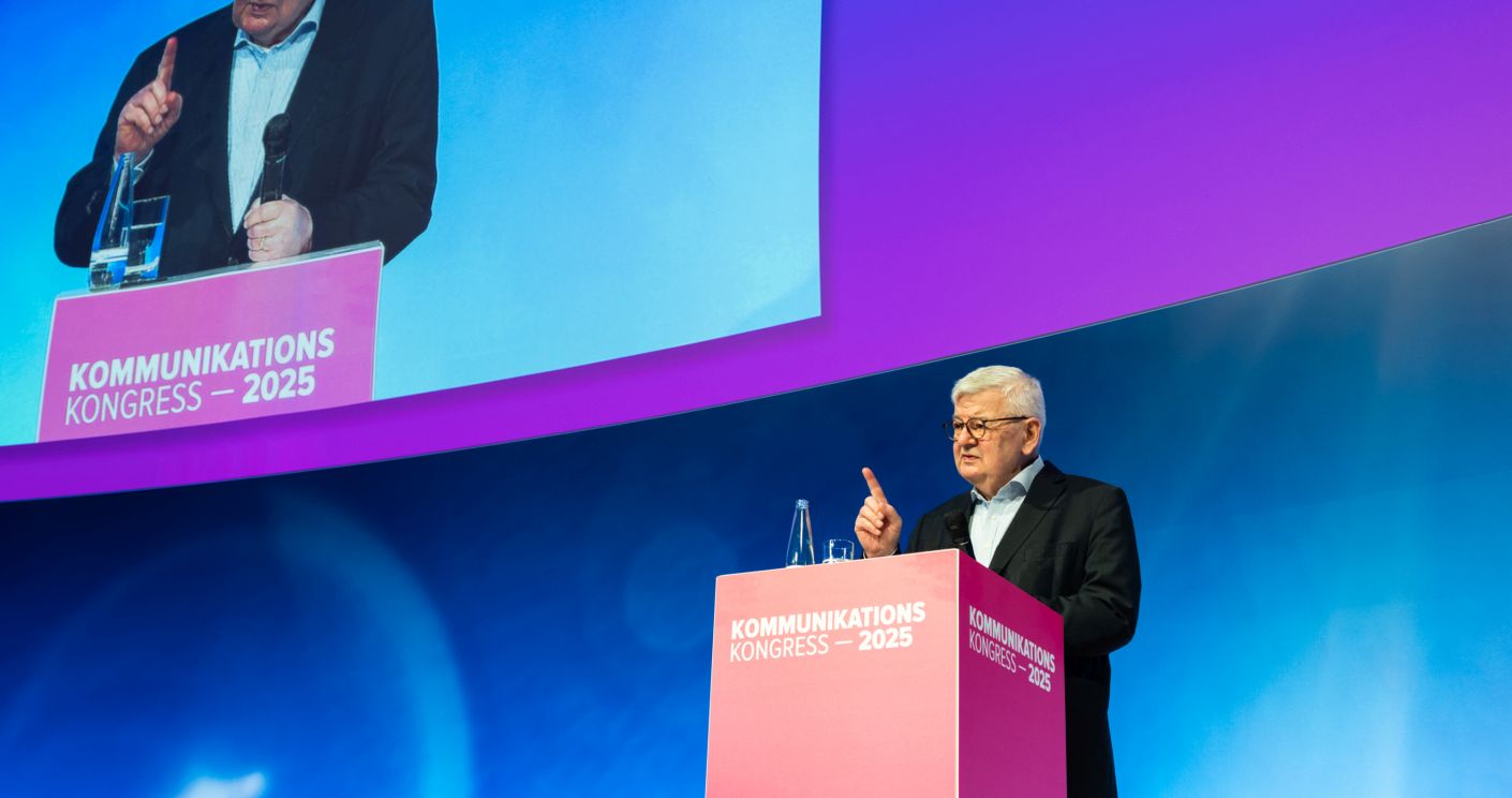 Joschka Fischer, älterer Mann mit weißen Haaren, Brille mit schwarzem Gestell und in schwarzem Anzug,, weißem Hemd und ohne Krawatte, spricht auf dem Kommunikationskongress 2025, Er hebt mahnend seinen Zeigefinger.
