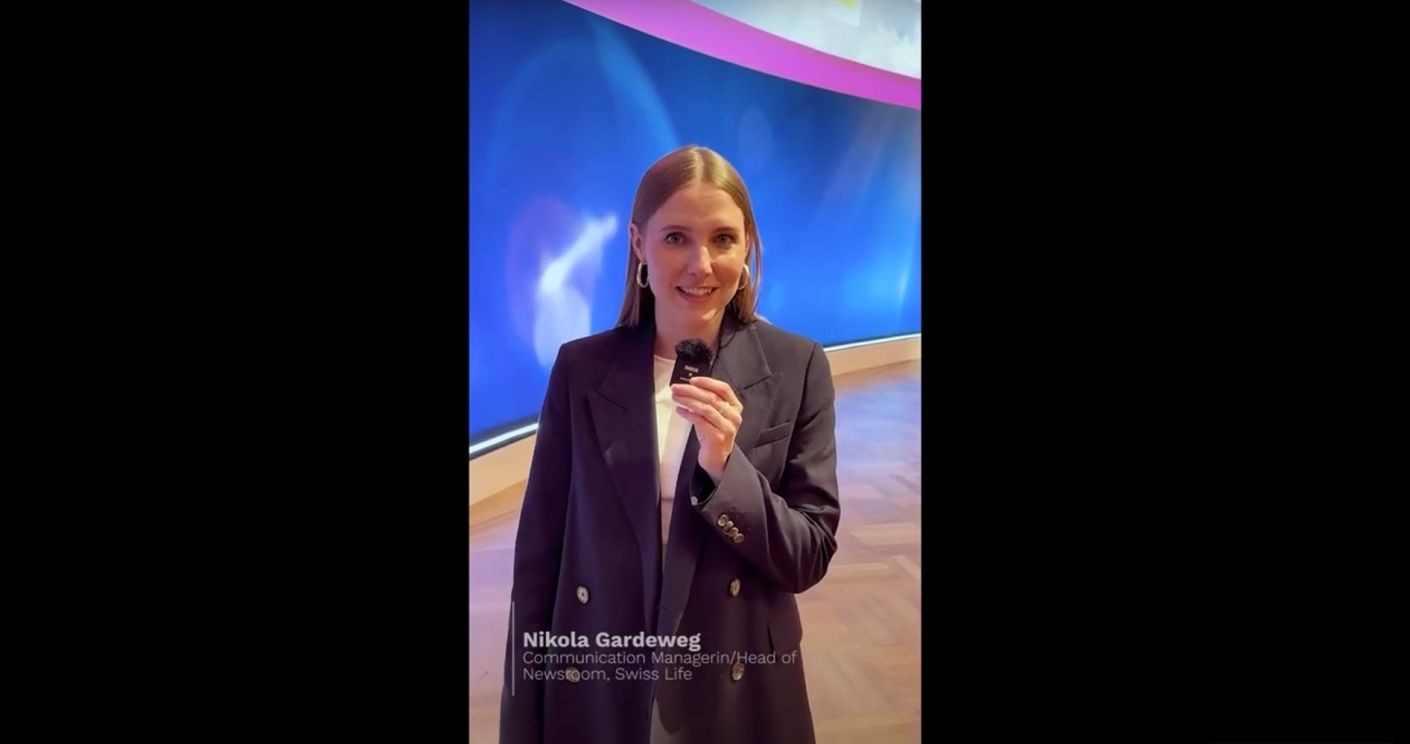 Frau mit Mikrofon in eleganter Kleidung vor einem bunten Hintergrund, spricht in die Kamera.