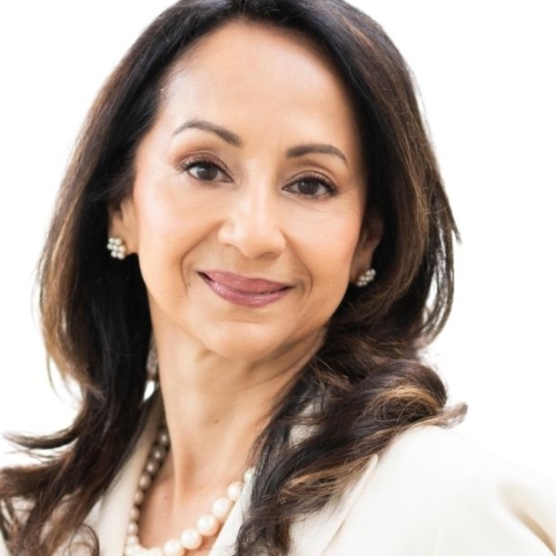 Porträt einer Frau mit langen, dunklen Haaren, elegantem Outfit und Perlenohrringen, lächelnd vor hellem Hintergrund.