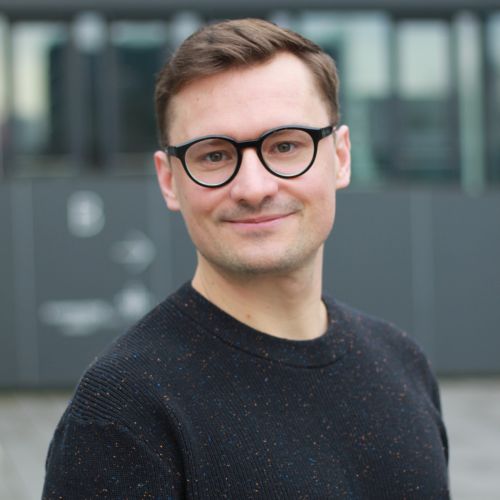 Mann mit Brille und dunklem Pullover lächelt vor einem modernen, unscharfen Hintergrund.