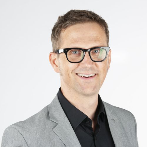 Porträt eines Mannes mit Brille, lächelnd, in einem grauen Anzug vor einem hellen Hintergrund.