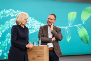 Zwei Personen stehen an einem kleinen Stehtisch, links eine Frau mit schulterlangen blond gefärbten Haaren in einem schwarzen Kostüm. Rechts von ihr ein Herr mit Brille, der sie anlächelt, den Kopf leicht auf die linke Hand gestützt, mit einem braun-melierten Jackett und dunkler Anzughose. Im Hintergrund eine Leinwand mit einem Pflanzenmotiv.