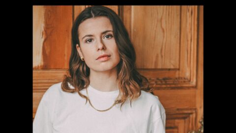 Porträt von Luisa Neubauer vor einer Holzwand. Neubauer trägt ihre Haare offen, dazu ein weißes T-Shirt, goldene Ohrringe und eine Goldkette.