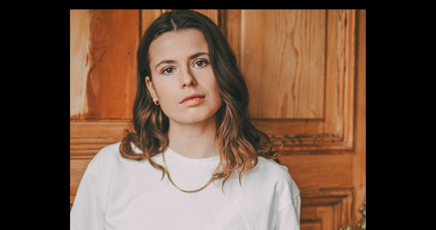 Porträt von Luisa Neubauer vor einer Holzwand. Neubauer trägt ihre Haare offen, dazu ein weißes T-Shirt, goldene Ohrringe und eine Goldkette.