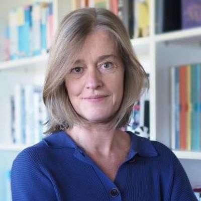 Porträt einer Frau mit blonden Haaren, die in einem blauen Pullover vor einem Bücherregal steht.