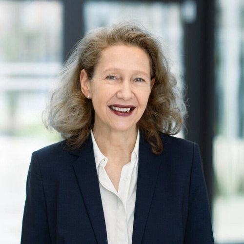 Porträt einer lächelnden Frau mit lockigem Haar, in einem dunklen Blazer und weißem Hemd, vor einem hellen Hintergrund.