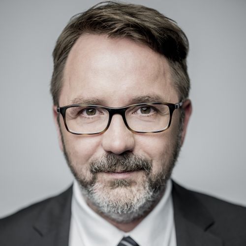 Porträt eines Mannes mit Brille, Bart und Anzug, vor einem neutralen Hintergrund.