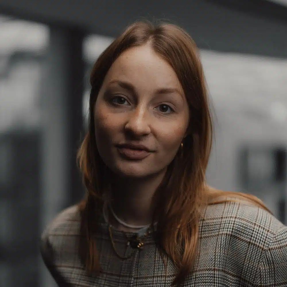 Porträt einer jungen Frau mit langen, roten Haaren, die in die Kamera schaut und einen karierten Blazer trägt.