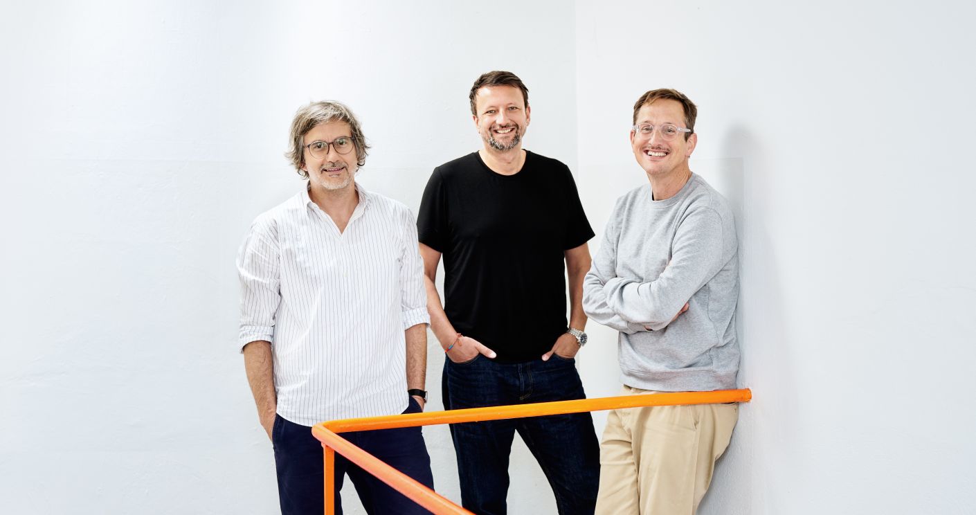 Drei Männer stehen lächelnd an einer orangefarbenen Geländer, vor einer weißen Wand.