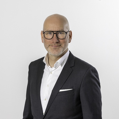 Porträt eines Mannes mit Glatze, Brille und Anzug, vor einem weißen Hintergrund.