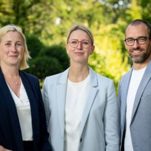 Drei Personen stehen im Freien: zwei Frauen in Blazern und ein Mann in einem hellen Anzug, mit grüner Natur im Hintergrund.