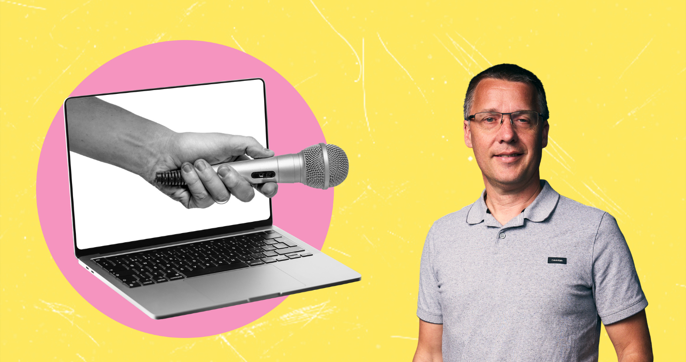 Mann mit Brille steht neben einem Laptop, auf dem eine Hand mit Mikrofon zu sehen ist, vor gelbem Hintergrund.