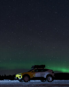 Ein Auto steht auf einer verschneiten Straße unter einem sternenklaren Himmel. Im Hintergrund sind grüne Polarlichter zu erkennen.