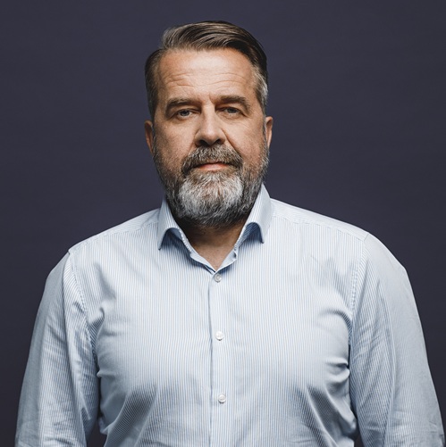 Porträt eines Mannes mit Bart, in einem hellblauen Hemd, vor einem dunklen Hintergrund.