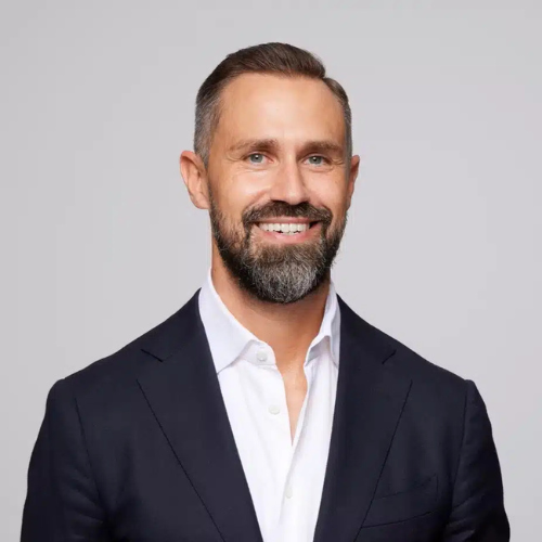 Porträt eines lächelnden Mannes mit Bart, in einem dunklen Anzug und weißem Hemd, vor neutralem Hintergrund.