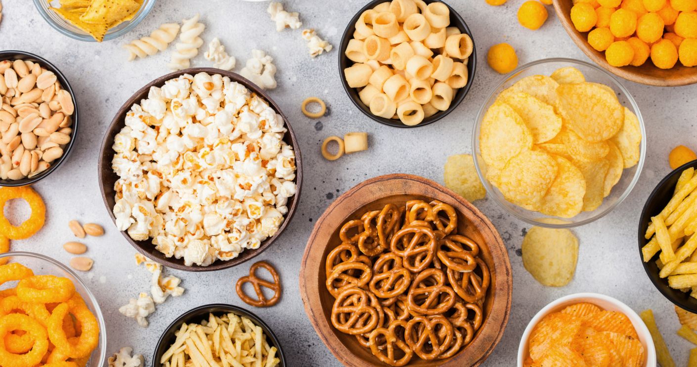 Bunte Auswahl an Snacks: Popcorn, Erdnüsse, Zwiebelringe, Brezeln, Chips und Käsebällchen in Schalen auf hellem Hintergrund.