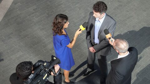 Politiker gibt ein Interview, umgeben von Reportern mit Mikrofonen und einer Kamera.