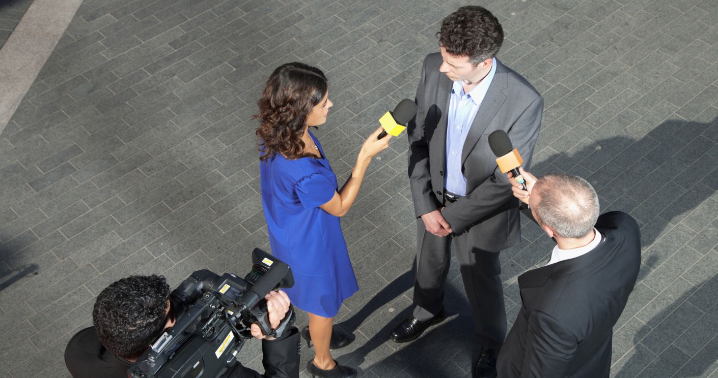 Politiker gibt ein Interview, umgeben von Reportern mit Mikrofonen und einer Kamera.