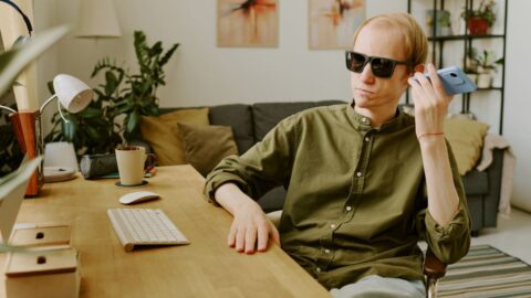 Mann mit schwarzer Sonnenbrille sitzt an einem Schreibtisch in einem wohnlichen Büro und hält sein Ohr an ein Handy.