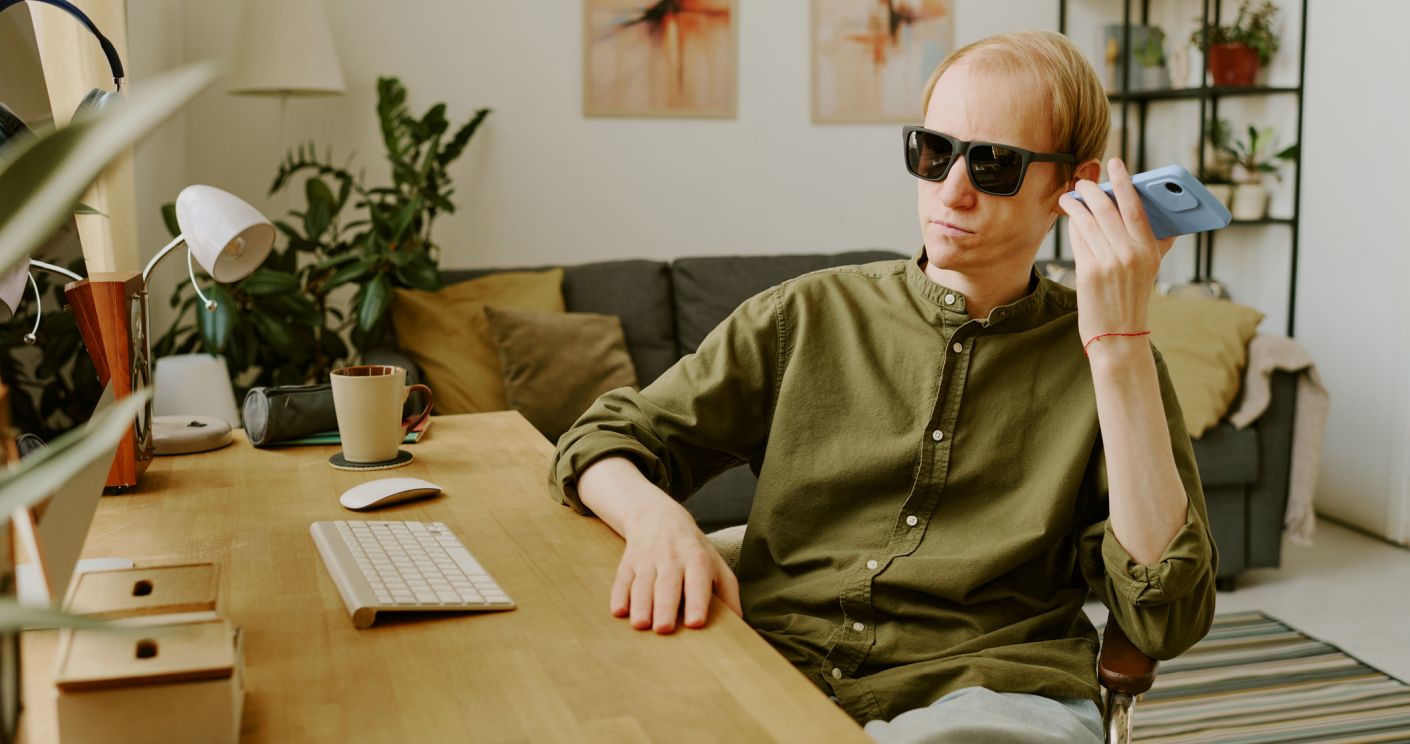 Mann mit schwarzer Sonnenbrille sitzt an einem Schreibtisch in einem wohnlichen Büro und hält sein Ohr an ein Handy.