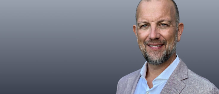 Porträt eines lächelnden Mannes mit Bart, in einem hellen Hemd und einem hellen Blazer, vor einem grauen Hintergrund.