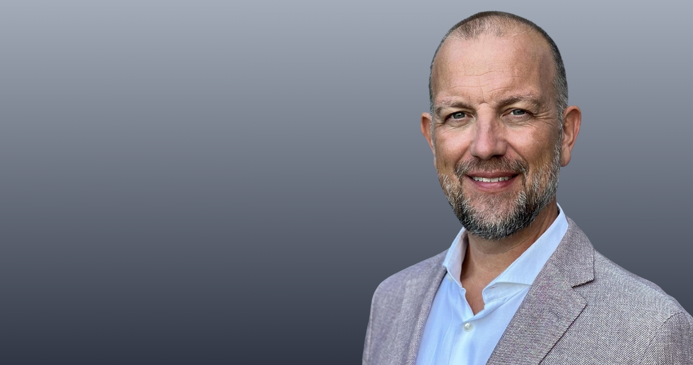 Porträt eines lächelnden Mannes mit Bart, in einem hellen Hemd und einem hellen Blazer, vor einem grauen Hintergrund.