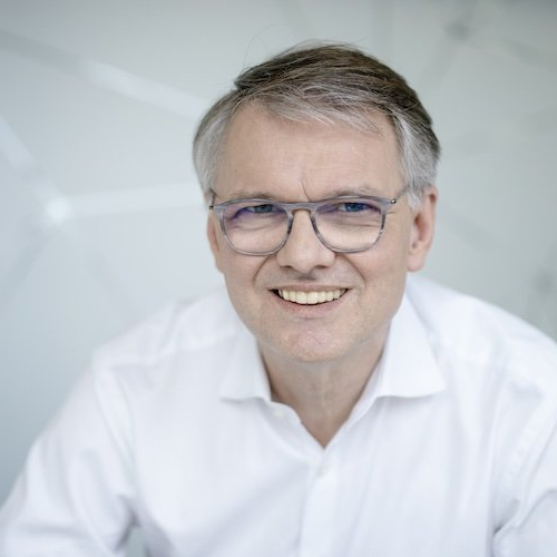 Porträt eines lächelnden Mannes mit Brille, in einem weißen Hemd vor einem hellen, modernen Hintergrund.