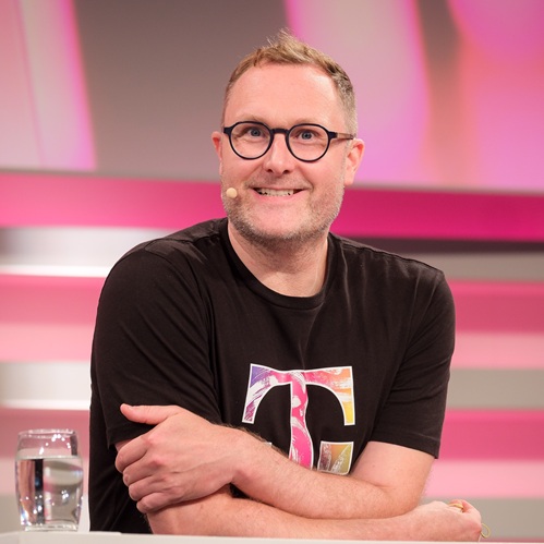 Mann mit Brille und T-Shirt mit Telekom-Logo lächelt am Tisch, im Hintergrund pinke Beleuchtung.