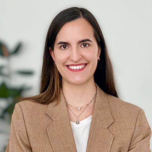 Porträt einer lächelnden Frau mit langen, braunen Haaren, die einen karierten Blazer trägt, vor einem neutralen Hintergrund.