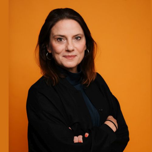 Porträt einer Frau mit braunen Haaren, die in einem schwarzen Oberteil vor einem orangefarbenen Hintergrund steht.