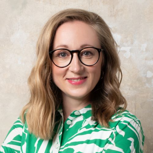Porträt einer Frau mit blonden, gewellten Haaren, Brille und grün-weißer Bluse vor einem neutralen Hintergrund.