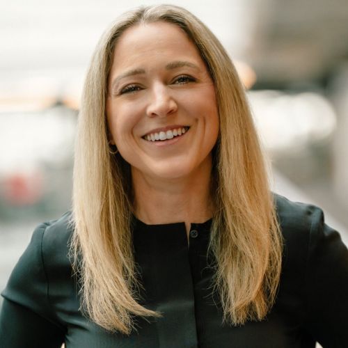 Frau mit langen, blonden Haaren und lächelndem Gesicht, trägt ein schwarzes Oberteil, steht in einem modernen Innenraum.