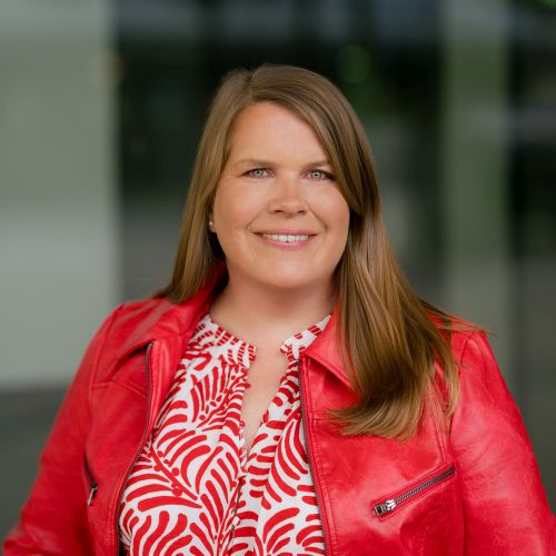 Porträt einer lächelnden Frau mit langen, braunen Haaren, in roter Lederjacke und gemusterter Bluse, vor unscharfem Hintergrund