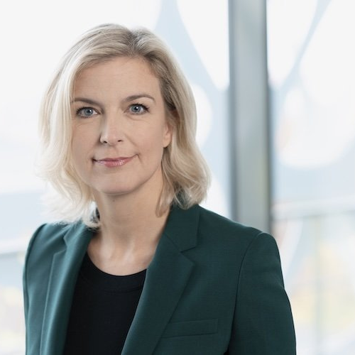 Porträt einer Frau mit blonden, schulterlangen Haaren, in einem grünen Blazer, vor einem modernen, hellen Hintergrund.