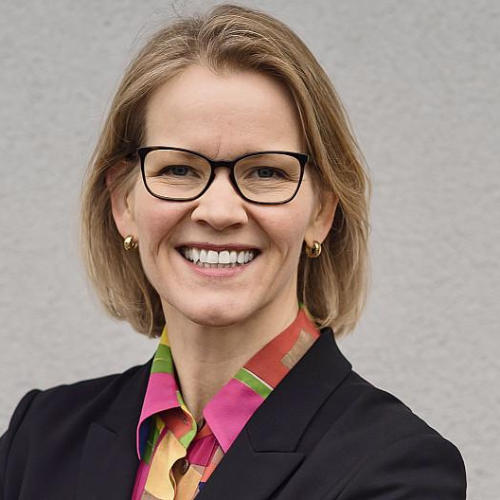 Porträt einer lächelnden Frau mit Brille, blonden Haaren, in einem schwarzen Blazer und buntem Hemd vor neutralem Hinter...