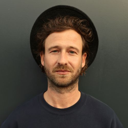 Mann mit lockigem Haar und Hut, trägt einen dunkelblauen Pullover, steht vor einer grauen Wand.