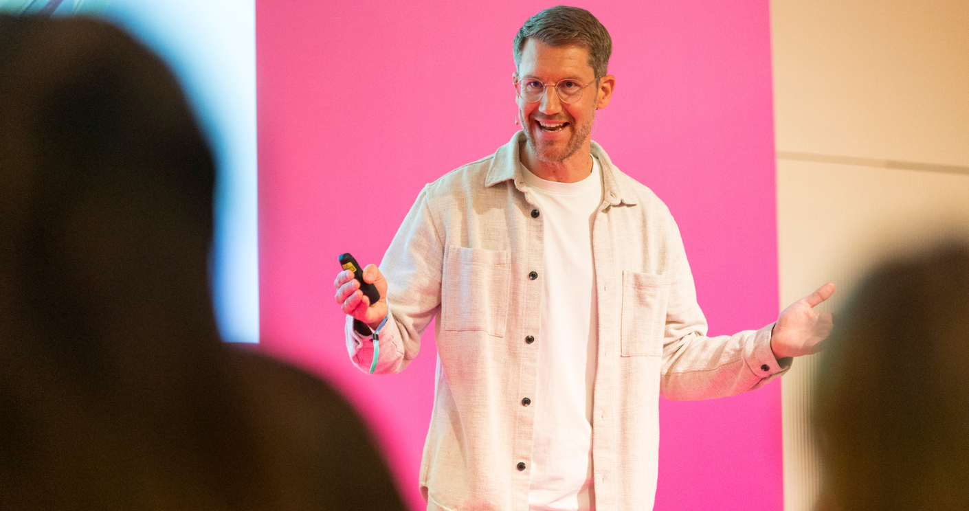 Mann mit Brille und lässiger Kleidung spricht auf einer Bühne vor einem pinken Hintergrund.