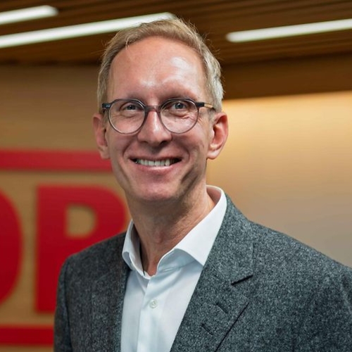 Porträt von Jens-Oliver Voß, Kommunikationschef der Deutschen Bahn, lächelnd vor einem modernen Hintergrund.