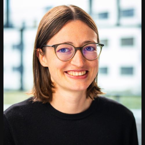 Porträt einer jungen Frau mit schulterlangen glatten braunen Haaren und Mittelscheitel, mit Brille, lächelnd vor einem unscharfen Hintergrund.