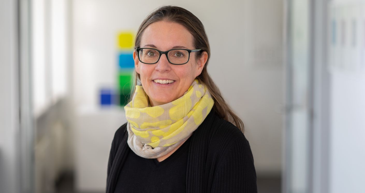 Porträt von Prof. Dr. Elke Kronewald mit Brille und gelbem Schal, lächelnd in einem modernen Büro.