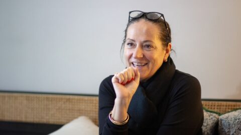 Frau mit einem schwarzen Pullover sitzt auf einem Sofa, gestikuliert mit ihrer Hand, die sie zu einer Faust geballt hat, gleichzeitig lächelt sie.