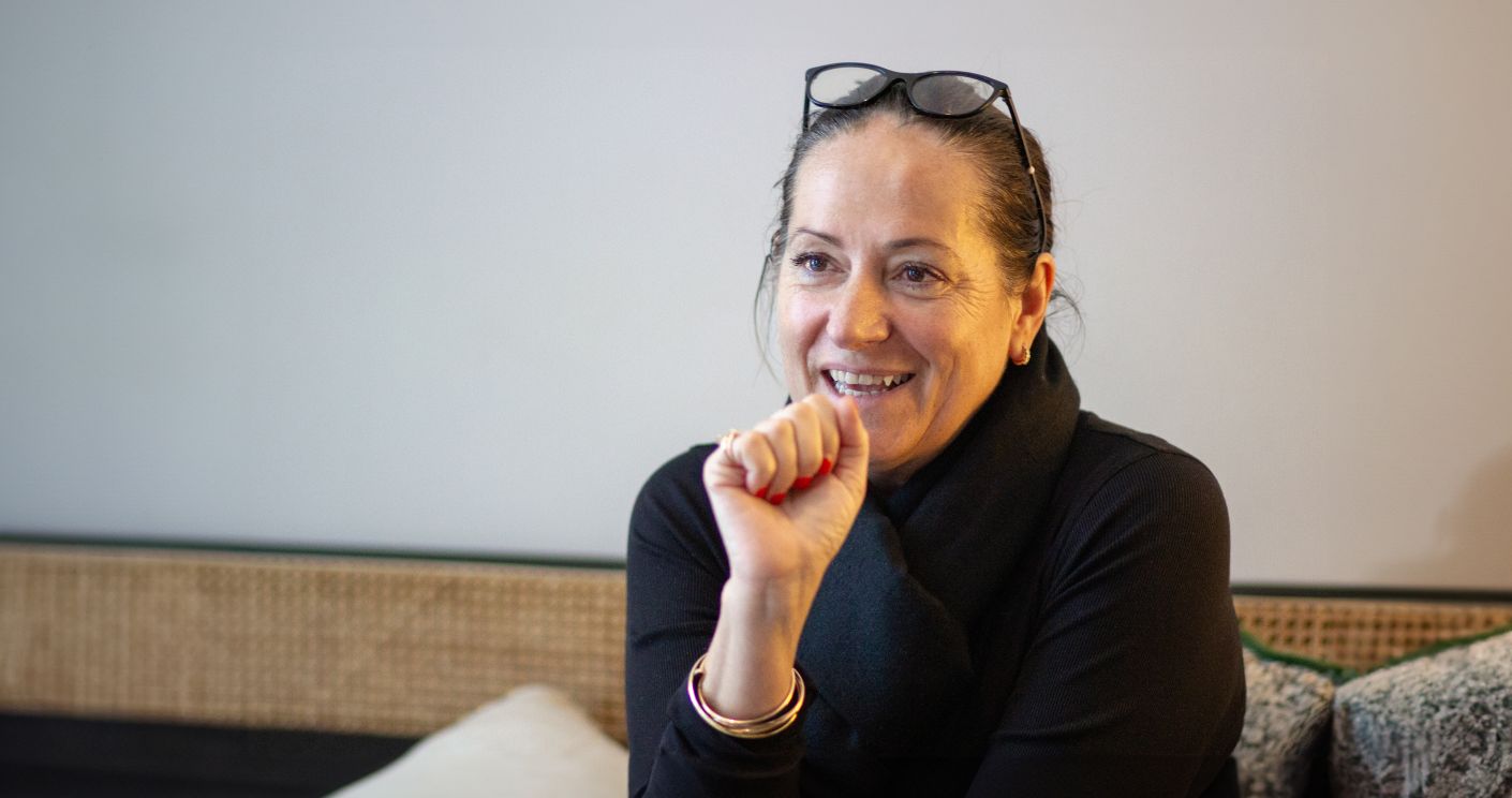 Frau mit einem schwarzen Pullover sitzt auf einem Sofa, gestikuliert mit ihrer Hand, die sie zu einer Faust geballt hat, gleichzeitig lächelt sie.