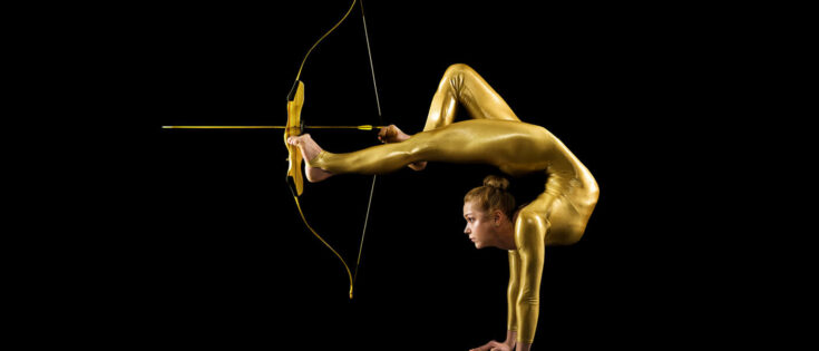 Akrobatin in goldenem Ganzkörperanzug vor schwarzem Hintergrund, die einen Handstand macht, mit ihren Füßen spannt sie einen Bogen, sie zielt.