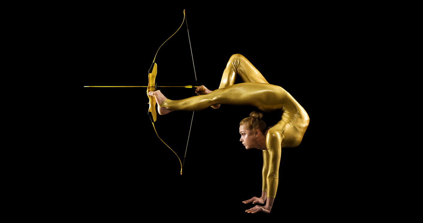 Akrobatin in goldenem Ganzkörperanzug vor schwarzem Hintergrund, die einen Handstand macht, mit ihren Füßen spannt sie einen Bogen, sie zielt.